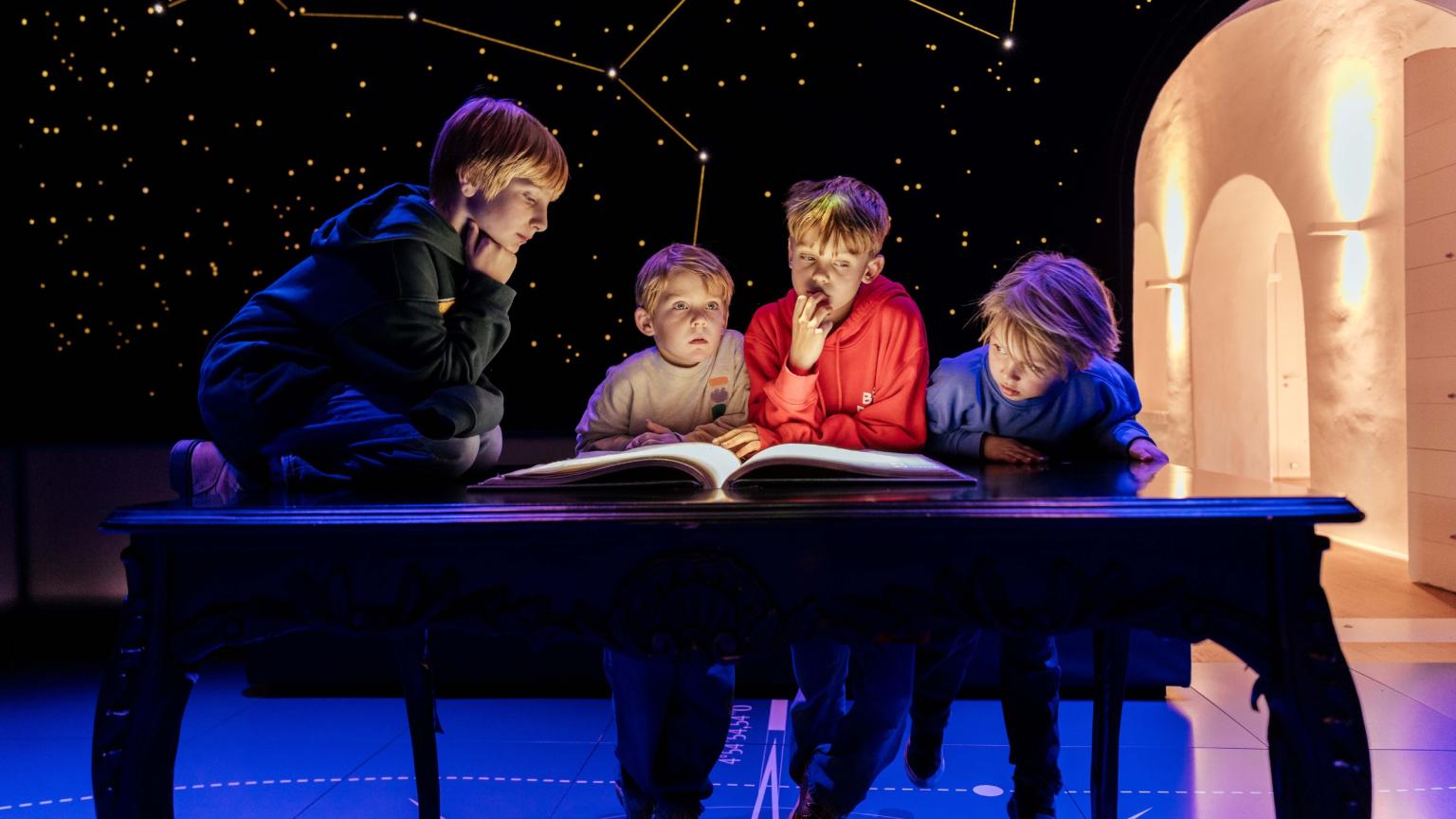 Groep kinderen leest samen in een groot boek onder een sterrenhemel-installatie in het museum.