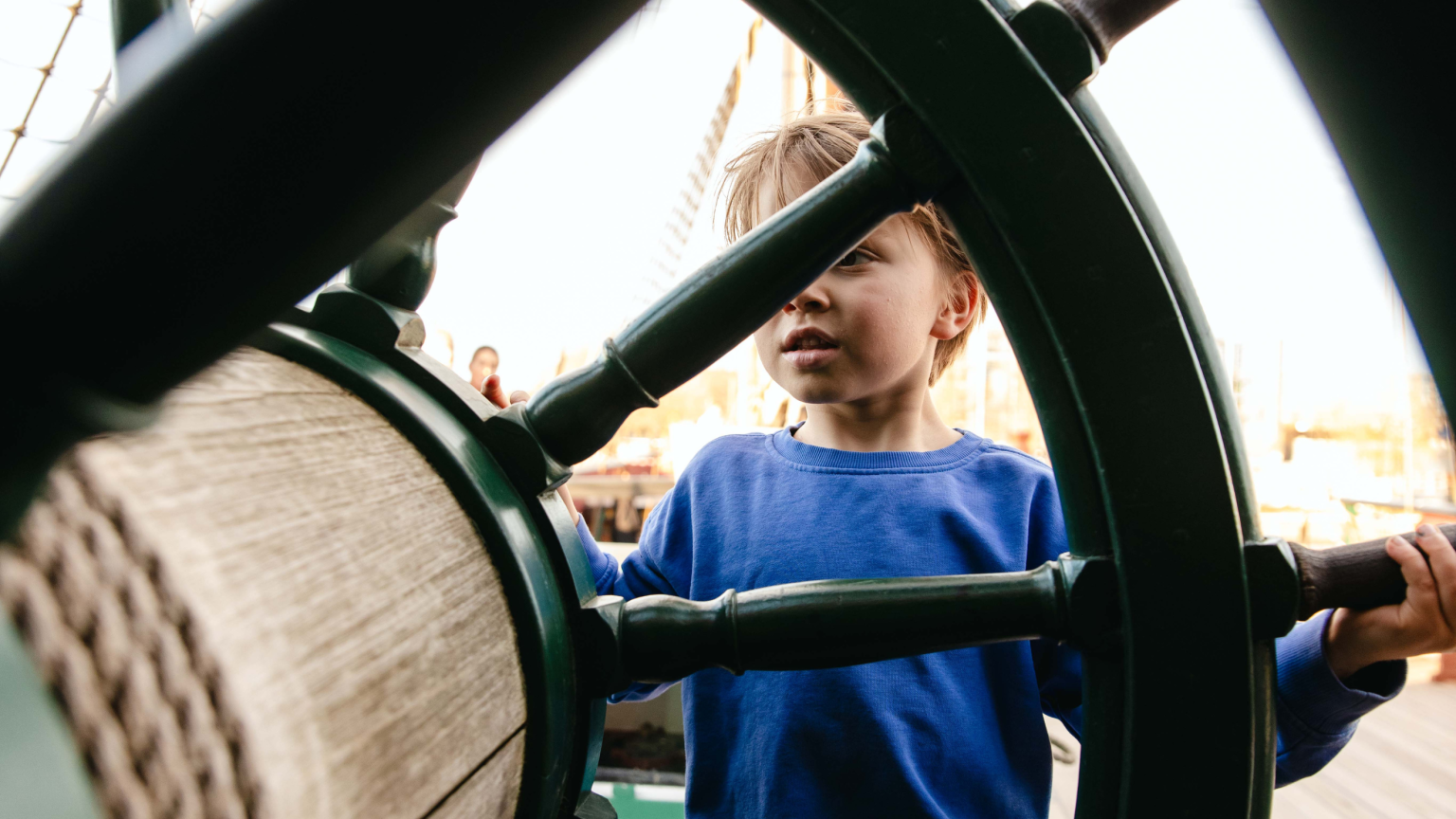 Kind dat het grote houten stuurwiel bedient aan boord van het VOC-schip Amsterdam.