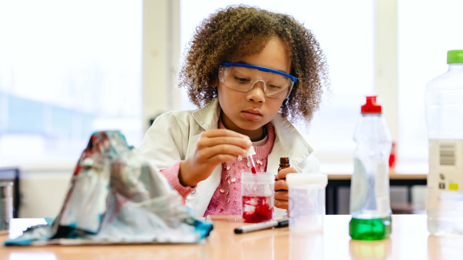Girl in lab coat and goggles performing a science experiment with red liquid at Hemlab