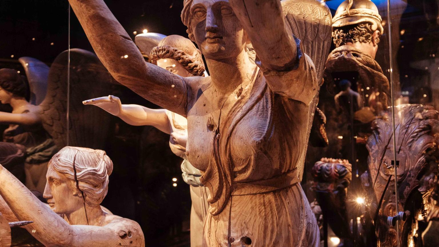 Houten beelden en scheepsornamenten tentoongesteld tegen een achtergrond van sterren en sterrenbeelden.