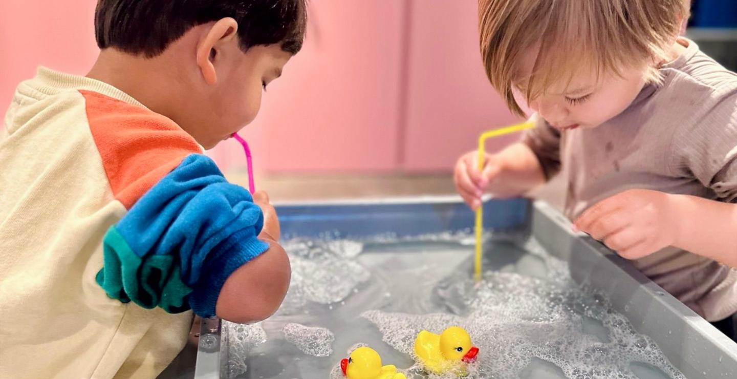 Kinderen spelen met water, schuim en gele badeendjes tijdens de Montessori-speelgroep.